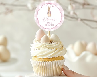 May include: A cupcake with white icing, decorated with three eggs and a round topper. The topper has a bunny illustration and the text "Olivia's Baby Shower" in pink, set against a pink gingham border.