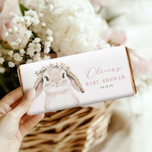 May include: A chocolate bar wrapper featuring a watercolor illustration of a bunny wearing a floral crown. The text "Olivia's BABY SHOWER 04.15.26" is printed in pink script. The wrapper is held in a person's hand.