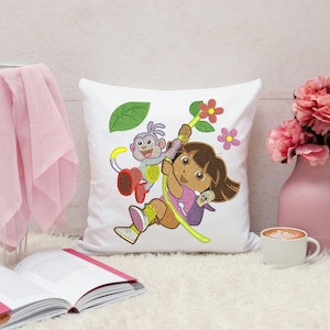 Puede incluir: Cojín cuadrado blanco con un diseño bordado de una niña de dibujos animados y un mono columpiándose en una liana. La niña tiene el pelo castaño y lleva una camisa rosa. El mono es gris y rojo. También hay flores rosas y naranjas y una hoja verde bordadas.