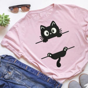 Op de afbeelding: Een lichtroze T-shirt met korte mouwen en een geborduurd zwart kattenontwerp. De kop en voorpoten van de kat verschijnen boven een horizontale lijn, terwijl de achterpoten en de staart onder een tweede horizontale lijn op de stof staan.