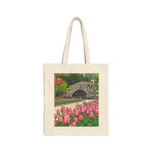 May include: Cream-colored tote bag featuring a colorful illustration of a stone bridge over a path lined with pink tulips. The scene includes trees and a small building in the background. The bag has two handles.