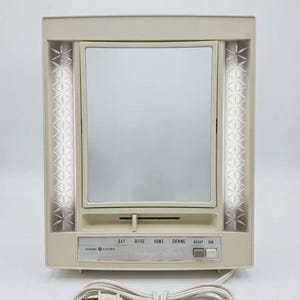 May include: A vintage cream-coloured General Electric makeup mirror. It features a rectangular mirror with vertical light strips. The base has settings for day, office, home, and evening lighting, with bright and dim options. A white power cord is attached.