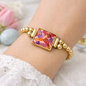 Puede incluir: Pulsera dorada con un elemento central cuadrado con un diseño abstracto colorido. La pulsera tiene un diseño de cuentas con esferas doradas y detalles rectangulares dorados. El elemento central presenta un patrón rojo, naranja, morado y azul.