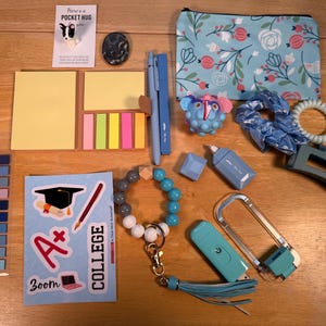 May include: A collection of school and office supplies, including sticky notes, pens, a floral print pouch, and a beaded bracelet. Other items include a blue stress ball, hair accessories, and a keychain with a tassel. The items are arranged on a wooden surface.