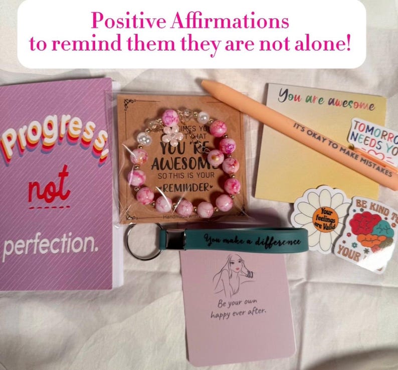 May include: An assortment of self-care items, including a pink and white beaded bracelet, a notebook with "Progress not perfection," and a pen with "It's okay to make mistakes." The image also includes stickers and a keychain.
