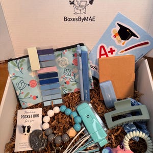 May include: An open gift box filled with various items, including a floral print pouch, pens, a hair clip, a keychain, and a beaded bracelet. The box also contains a sticker with a graduation cap and the text "A+". The box is labeled "BoxesByMAE".