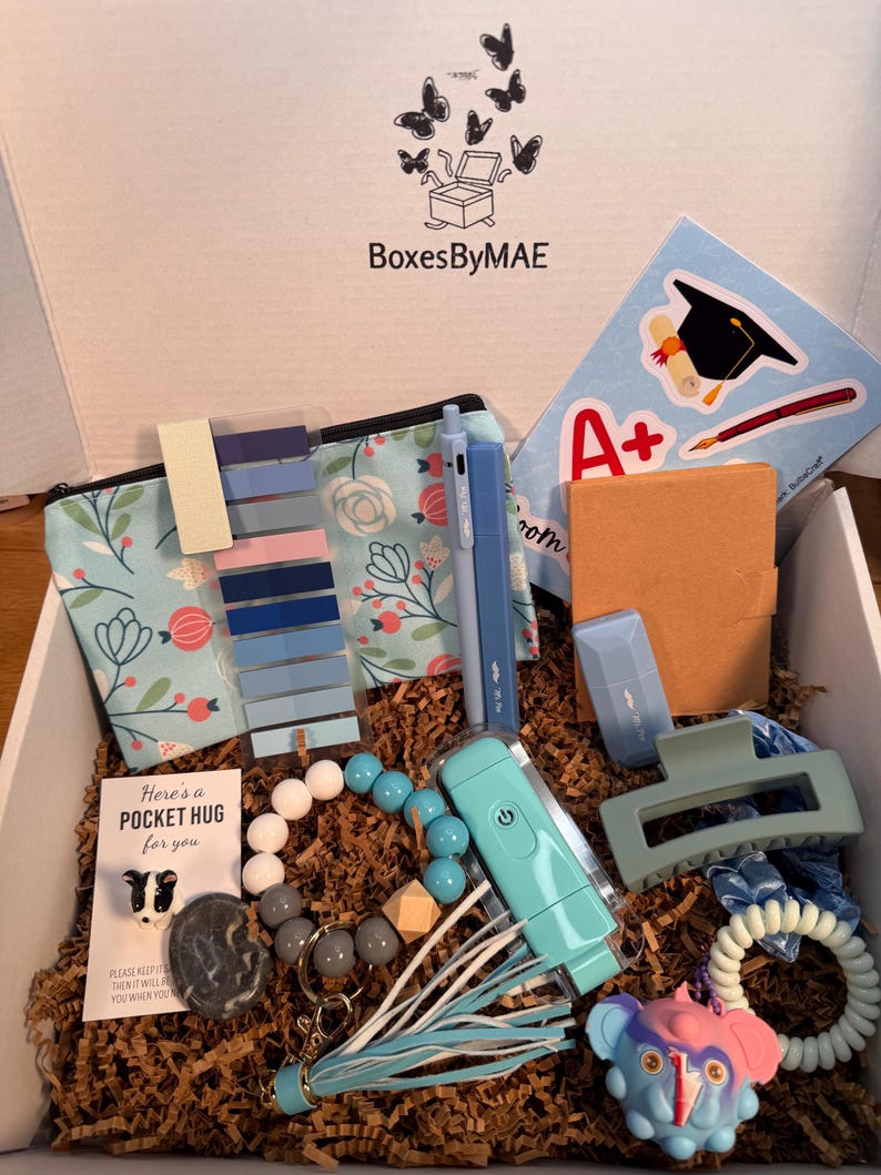 May include: An open gift box filled with various items, including a floral print pouch, pens, a hair clip, a keychain, and a beaded bracelet. The box also contains a sticker with a graduation cap and the text "A+". The box is labeled "BoxesByMAE".