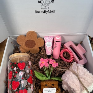 May include: A gift box containing a glass cup with strawberry prints and a bamboo lid, a flower-shaped cork coaster, a tube of cherry blossom hand cream, a pink spray bottle, a pink hair claw, a pink coil hair tie, a crochet plant, and fuzzy socks.
