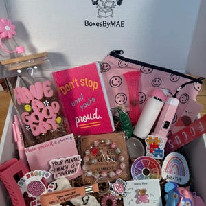May include: An open box filled with pink and white self-care items. Includes a water bottle, journal, stickers, bracelet, scrunchie, and a pink cosmetic bag with smiley faces. The box has the text "BoxesByMAE".