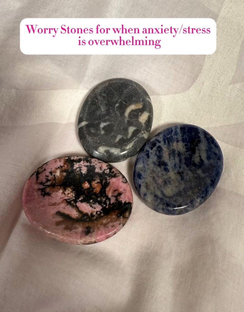May include: Three smooth worry stones in shades of pink, black, gray, and blue. The stones are oval-shaped and polished, with unique patterns and textures. Text on the image reads: Worry Stones for when anxiety/stress is overwhelming.