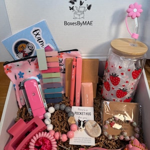 May include: A gift box filled with pink and gray accessories, including a hair clip, scrunchie, pens, and a beaded keychain. A glass jar with a strawberry design and a flower-shaped straw is also visible. The box has the text "BoxesByMAE".