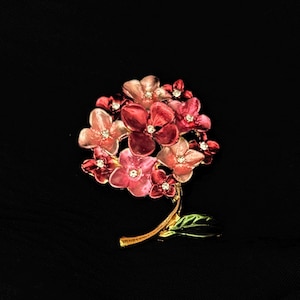 Könnte beinhalten: Blumenbrosche mit roten und rosa Blüten und kleinen glitzernden Akzenten. Die Brosche hat einen goldfarbenen Stiel und ein grünes Blatt, vor schwarzem Hintergrund.