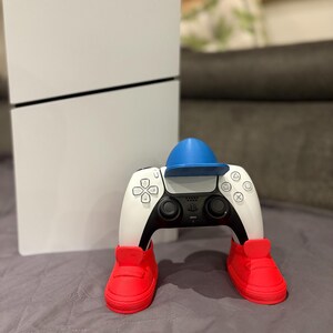 May include: A white PlayStation 5 console is positioned next to a white and black controller wearing a blue hat and red shoes. The controller has a black touchpad and buttons. The console has a black stripe.