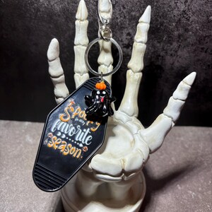 May include: A black motel-style keyring with the text "Spooky is my favourite season" in orange and white lettering. The keyring features a small black ghost charm with an orange pumpkin on its head, attached to a silver metal ring and clasp.