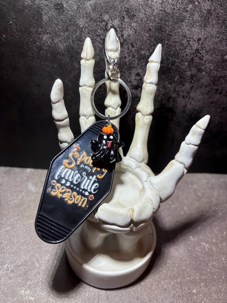 May include: A black motel-style keyring with the text "Spooky is my favourite season" in orange and white lettering. The keyring features a small black ghost charm with an orange pumpkin on its head, attached to a silver metal ring and clasp.