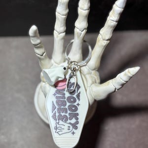 May include: A white motel-style keyring with the text SPOOKY VIBES in glittery black lettering, featuring small ghost illustrations and a pink circle accent. The keyring includes a silver metal ring and a small white ghost charm attached.