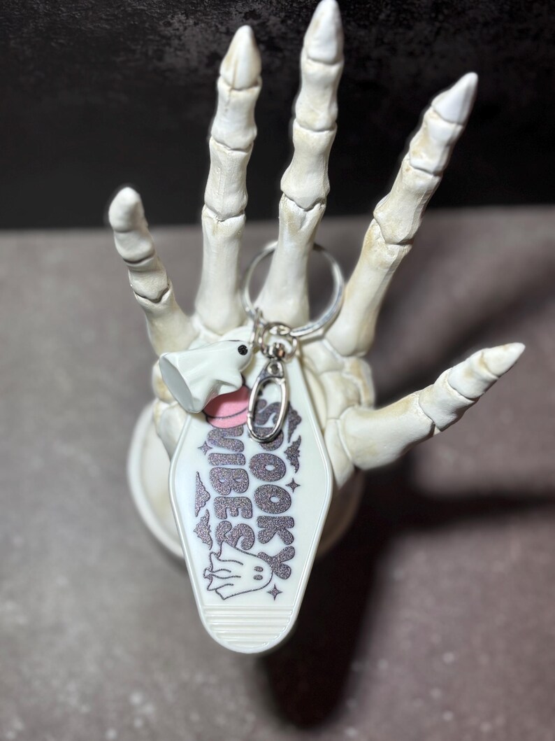 May include: A white motel-style keyring with the text SPOOKY VIBES in glittery black lettering, featuring small ghost illustrations and a pink circle accent. The keyring includes a silver metal ring and a small white ghost charm attached.