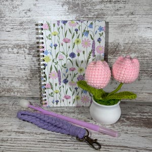 May include: A spiral-bound notebook with a floral design, a pair of pink crochet tulips in a white pot, a purple macrame wristlet with a clasp, and a purple pen with a white tip.