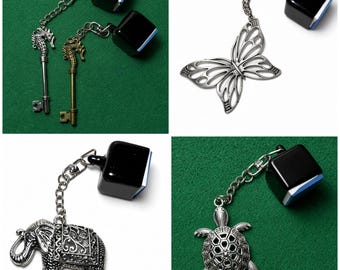 May include: A collection of four keychains featuring silver and bronze seahorse keys, a silver butterfly, a silver elephant, and a silver turtle. Each charm is attached by a metal chain to a black square block with a blue base accent.