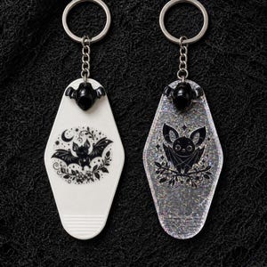 May include: Two motel-style keychains with silver-tone metal rings and chains. One is solid white with a black graphic of a bat, moon, and flowers. The other is clear with silver glitter, featuring a black graphic of a bat perched on branches.