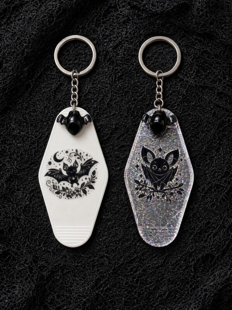 May include: Two motel-style keychains with silver-tone metal rings and chains. One is solid white with a black graphic of a bat, moon, and flowers. The other is clear with silver glitter, featuring a black graphic of a bat perched on branches.