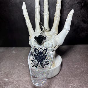 May include: A motel-style keyring made of clear resin with embedded silver glitter. The keyring features a black graphic illustration of a bat perched on flowers, a small black bat-shaped charm at the top, and a silver metal split ring attachment.
