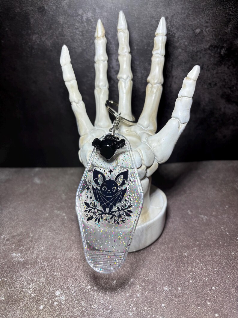 May include: A motel-style keyring made of clear resin with embedded silver glitter. The keyring features a black graphic illustration of a bat perched on flowers, a small black bat-shaped charm at the top, and a silver metal split ring attachment.