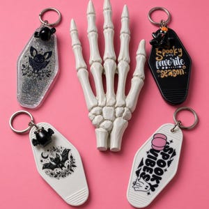 May include: Four coffin-shaped keychains with metal rings and a central white skeleton hand model. One keychain is clear with glitter and a bat, one is black with text reading Spooky is my favourite season, one is white with a bat, and one is white with text reading Spooky Vibes.