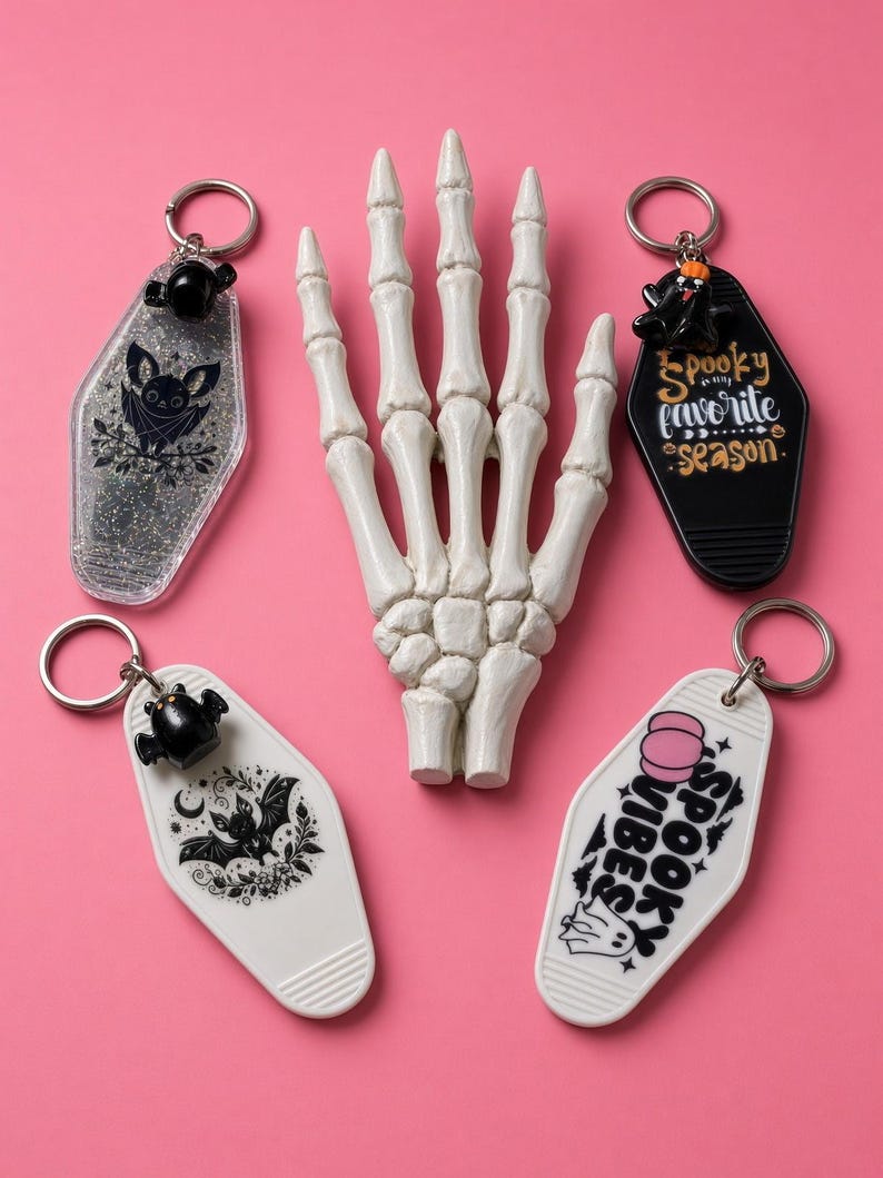 May include: Four coffin-shaped keychains with metal rings and a central white skeleton hand model. One keychain is clear with glitter and a bat, one is black with text reading Spooky is my favourite season, one is white with a bat, and one is white with text reading Spooky Vibes.