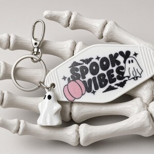 May include: A white motel-style keyring featuring the black text "SPOOKY VIBES," a pink pumpkin graphic, and small bat illustrations. It includes a silver metal ring with a small white ghost charm attached to the side of the keyring.