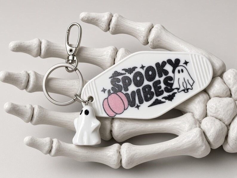 May include: A white motel-style keyring featuring the black text "SPOOKY VIBES," a pink pumpkin graphic, and small bat illustrations. It includes a silver metal ring with a small white ghost charm attached to the side of the keyring.