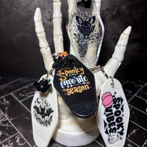 May include: Three coffin-shaped keychains with metal rings. One is clear with holographic glitter and a bat illustration. Another is black with the text "Spooky is my favourite season." The third is white with a pink pumpkin and "SPOOKY VIBES" text.