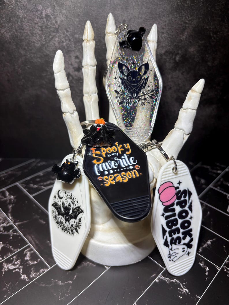 May include: Three coffin-shaped keychains with metal rings. One is clear with holographic glitter and a bat illustration. Another is black with the text "Spooky is my favourite season." The third is white with a pink pumpkin and "SPOOKY VIBES" text.