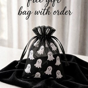 May include: A black sheer organza drawstring gift bag featuring a repeating pattern of small white ghosts with smiling faces. The text free gift bag with order is printed in black cursive script above the bag.