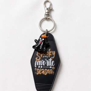 May include: A black motel-style keyring featuring a small black ghost charm with an orange pumpkin on its head. The keyring is printed with the white and orange text "spooky is my favorite season" and includes a silver metal lobster clasp and ring.