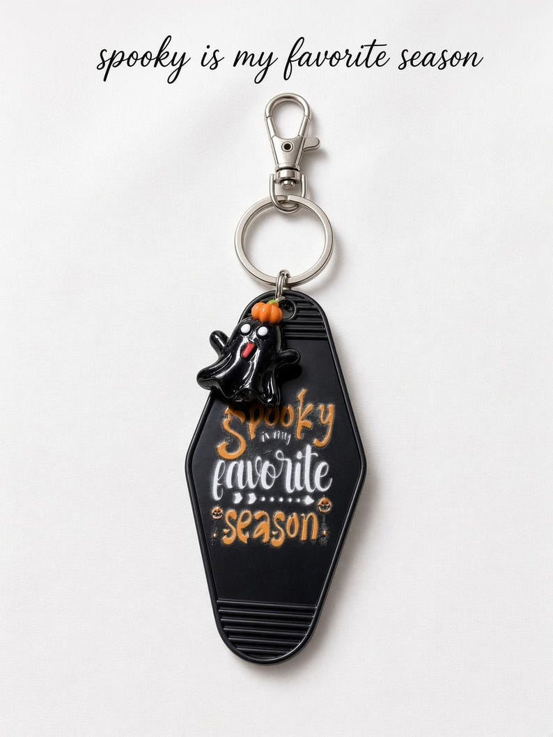 May include: A black motel-style keyring featuring a small black ghost charm with an orange pumpkin on its head. The keyring is printed with the white and orange text "spooky is my favorite season" and includes a silver metal lobster clasp and ring.