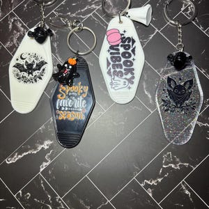 May include: Four motel-style keychains with metal rings. One white with a black bat, one black with a ghost and the text Spooky is my favourite season, one white with a pink pumpkin and the text SPOOKY VIBES, and one clear glittery keychain with a black bat.