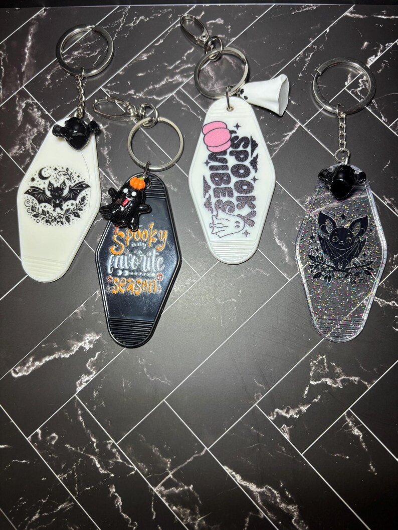 May include: Four motel-style keychains with metal rings. One white with a black bat, one black with a ghost and the text Spooky is my favourite season, one white with a pink pumpkin and the text SPOOKY VIBES, and one clear glittery keychain with a black bat.