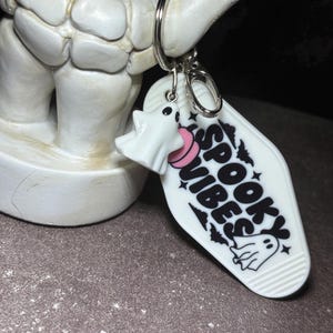 May include: A white motel-style keyring featuring the black text SPOOKY VIBES, a small ghost illustration, and a pink circular accent. A separate white ghost charm is attached to the silver metal ring, which is held by a skeletal hand figure.