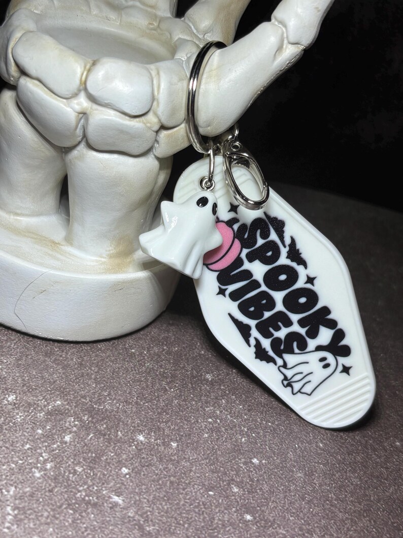 May include: A white motel-style keyring featuring the black text SPOOKY VIBES, a small ghost illustration, and a pink circular accent. A separate white ghost charm is attached to the silver metal ring, which is held by a skeletal hand figure.