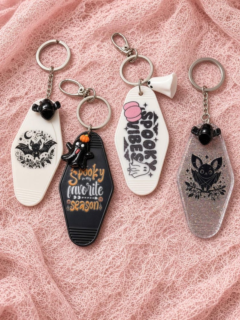 May include: Four motel-style keyrings with metal rings. One white with a black bat, one black with a ghost and the text 'Spooky is my favorite season', one white with a pink pumpkin and 'SPOOKY VIBES', and one clear glittery keyring with a black bat.