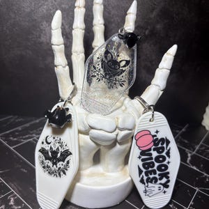 May include: Three coffin-shaped keychains featuring bat and ghost illustrations. One clear keychain has glitter and a bat, one white keychain shows a bat with floral accents, and one white keychain displays the text SPOOKY VIBES with a pink pumpkin.