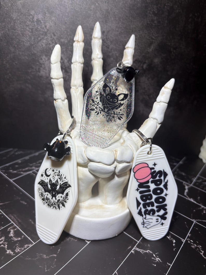 May include: Three coffin-shaped keychains featuring bat and ghost illustrations. One clear keychain has glitter and a bat, one white keychain shows a bat with floral accents, and one white keychain displays the text SPOOKY VIBES with a pink pumpkin.