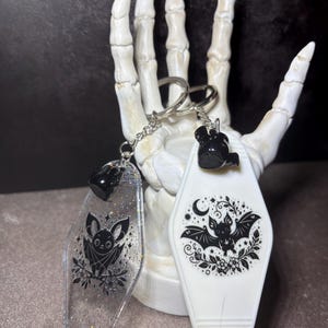 May include: Two motel-style keychains featuring bat illustrations. One is clear with silver glitter and a black bat graphic, while the other is opaque white with a black bat, moon, and floral design. Both have silver metal rings and chains.