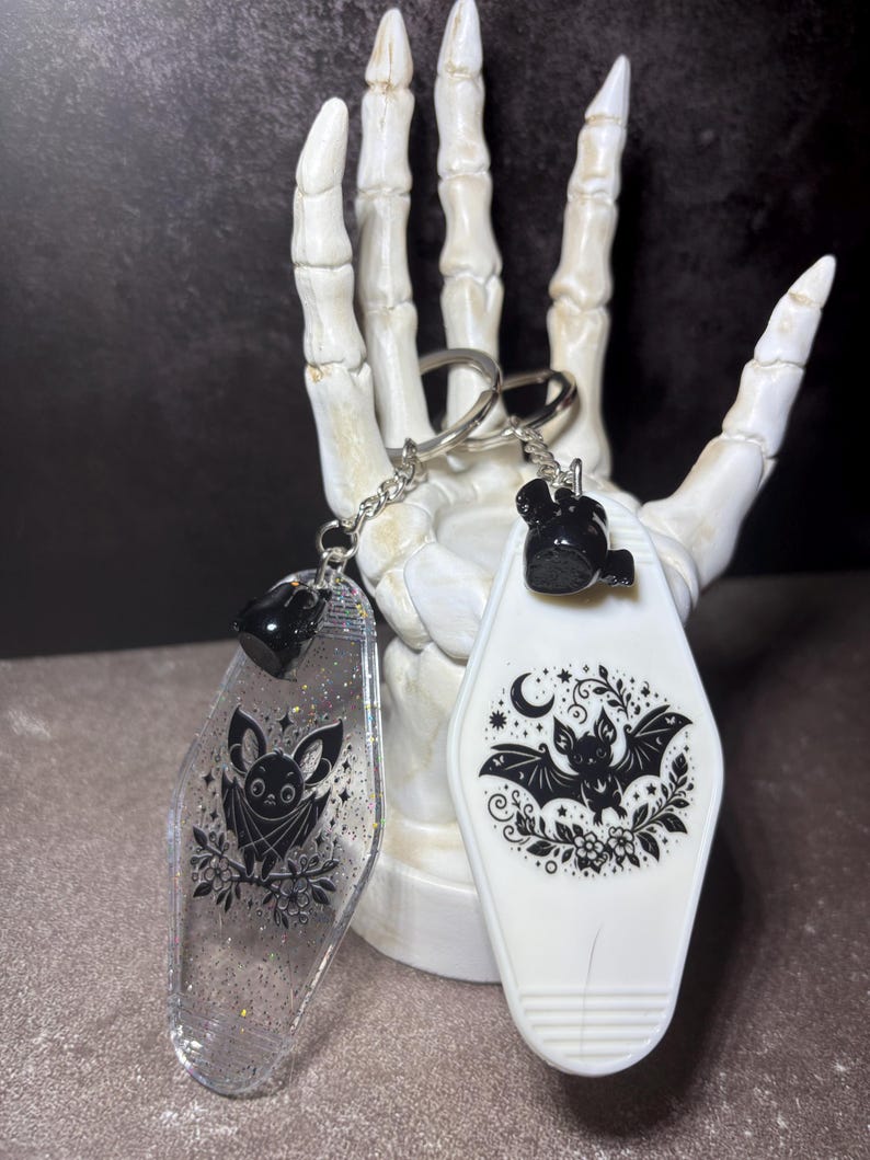 May include: Two motel-style keychains featuring bat illustrations. One is clear with silver glitter and a black bat graphic, while the other is opaque white with a black bat, moon, and floral design. Both have silver metal rings and chains.