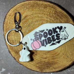 May include: A white motel-style keyring featuring the black glitter text SPOOKY VIBES, a small pink pumpkin, and a ghost illustration. A silver metal ring and lobster clasp are attached, along with a small white ghost charm figurine.