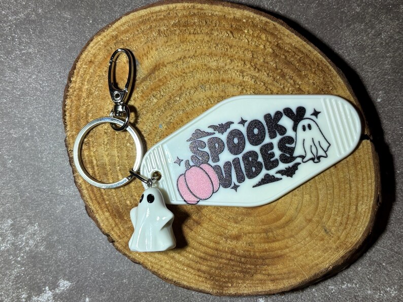 May include: A white motel-style keyring featuring the black glitter text SPOOKY VIBES, a small pink pumpkin, and a ghost illustration. A silver metal ring and lobster clasp are attached, along with a small white ghost charm figurine.