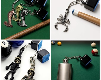 May include: A collection of billiard chalk holders featuring various metal charms including cobra snakes, a scorpion, and skull figures, attached to black or camouflage paracord and metal chains, paired with square blue billiard chalk cubes.