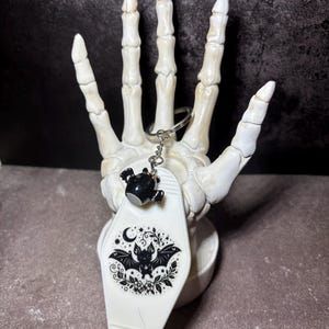 May include: A white motel-style keyring featuring a black graphic of a bat surrounded by stars, moons, and floral elements. The keyring includes a small black bat charm attached to a silver metal ring and chain, displaying a detailed illustration.