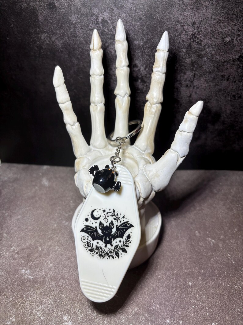 May include: A white motel-style keyring featuring a black graphic of a bat surrounded by stars, moons, and floral elements. The keyring includes a small black bat charm attached to a silver metal ring and chain, displaying a detailed illustration.